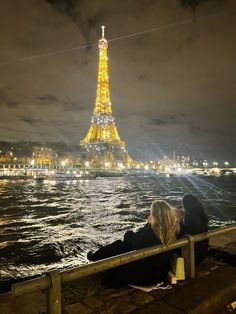 two people sitting on a bench in front of the eiffel tower at night