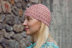 a woman wearing a pink crochet hat looks off into the distance while standing in front of a stone wall