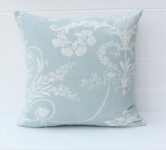 a light blue pillow with white flowers and vines on the front, against a white brick wall