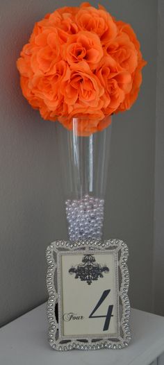 an orange flower in a clear vase on top of a white table with a silver frame