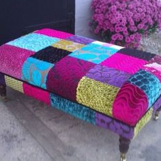 a patchwork bench sitting on the sidewalk next to some purple and pink flowers in front of a building