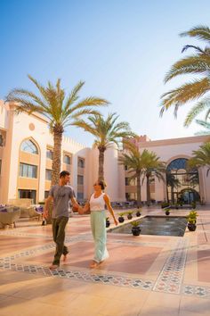 a man and woman holding hands in front of a building with palm trees on the ground