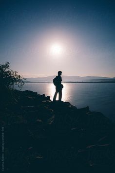 a man standing on the edge of a cliff looking out at the water under a full moon