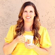 a woman holding a coffee cup in her right hand and smiling while standing against a wall