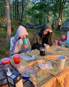 two people sitting at a picnic table in the woods drinking coffee and talking to each other