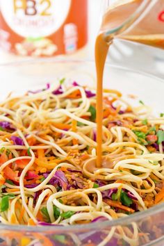 someone pouring dressing into a bowl filled with noodles and veggies, including carrots