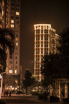 people are walking down the street at night in front of tall buildings with lights on them