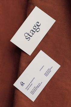 two white business cards sitting on top of a brown velvet material covered in black ink