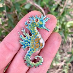 a small blue and yellow seahorse brooch in someone's hand with green grass behind it