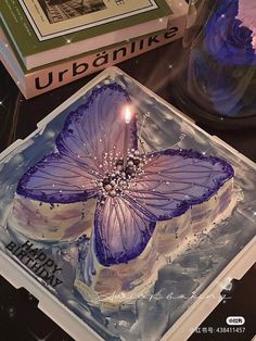 a birthday cake with a purple butterfly on it sitting next to a book and candle