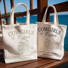 two bags sitting on top of a wooden table next to the ocean with cowgirls and geniuses logo