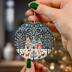 a woman holding up a christmas ornament in front of a tree with houses and deer