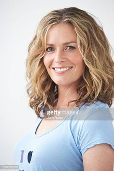 a woman with blonde hair and blue shirt smiling in front of a white background, looking at the camera