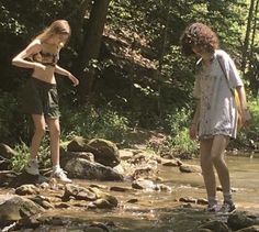 two young women standing on rocks in the water near some trees and rocks, while one looks at her cell phone