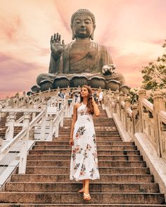 a woman in a white dress standing at the top of stairs with a large buddha statue behind her