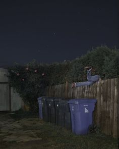 a man laying on top of a wooden fence next to trash cans in the yard