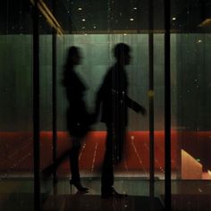 two people are walking in front of a glass wall with the reflection of lights on it