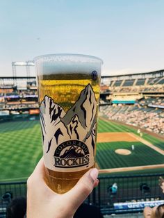 a person holding up a beer in front of a baseball field with mountains on it