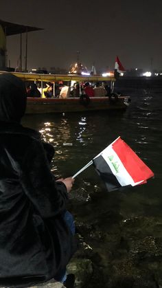 a person in a hooded jacket holding an egyptian flag on the edge of a body of water