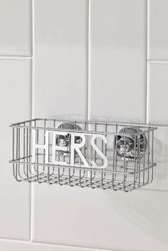 there is a metal basket on the wall with two hair dryers in it that says hers