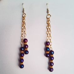 two pairs of gold and purple beaded earrings on a white surface, one is dangling from a chain