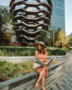 a woman in a hat sitting on a wall reading a paper and looking at the camera