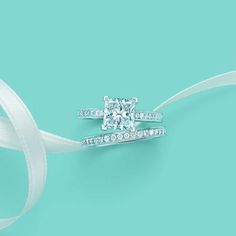 two wedding rings with diamonds on top and white ribbon around the band, against a turquoise background