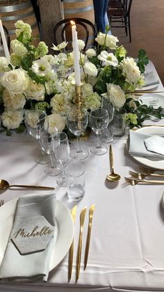 the table is set with white flowers and gold place settings, silverware and candlesticks