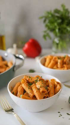 two white bowls filled with pasta next to silverware and tomatoes on a table in the background