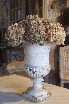 a white vase filled with lots of flowers on top of a wooden table next to a chair