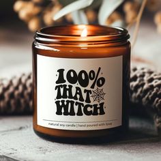 a candle that says 100 % that witch next to some pine cones and a potted plant