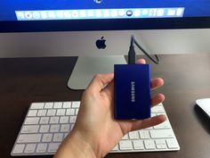 a hand is holding an external hard drive in front of a computer monitor and keyboard