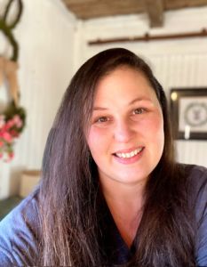 a woman with long brown hair smiling at the camera and wearing a blue shirt in front of a fireplace