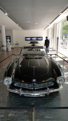 an old black car is parked in a museum with people looking at the cars on display