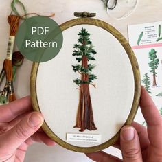 a person holding up a cross stitch pattern in front of an embroidery hoop with trees on it