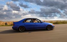 a blue car driving down the road with cloudy skies in the backgrouund