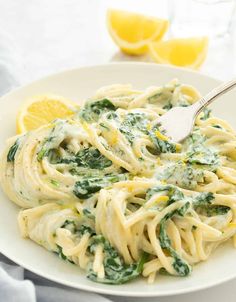 a white plate topped with pasta covered in spinach and sauce next to lemon wedges