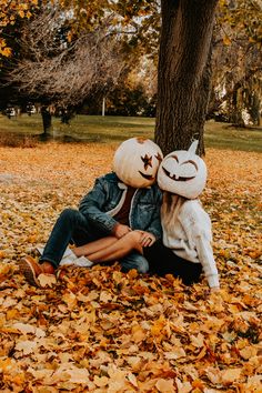 two people sitting under a tree with leaves on the ground and one person wearing a pumpkin mask