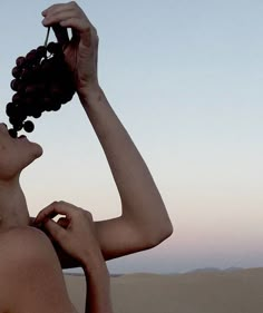 a woman holding grapes up to her face while standing in the desert at sunset or dawn