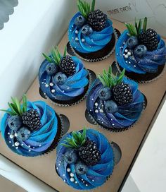 four cupcakes with blue icing and blackberries on top in a box