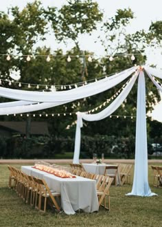 an outdoor wedding setup with white draping and wooden chairs, table cloth draped over it