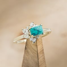 a gold ring with a turquoise stone surrounded by white diamonds on top of a wooden stand