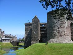 an old castle sitting on top of a lush green field next to a river in front of tall buildings