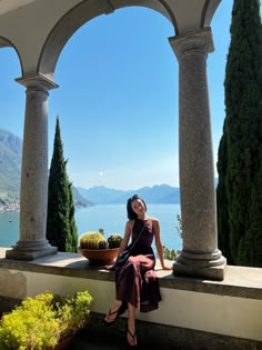 a woman in a dress sitting on a stone wall next to some trees and water