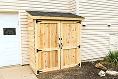 a wooden storage shed sitting in front of a white building with two doors and a black roof