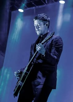 a man in a black suit holding a guitar on stage with blue lighting behind him