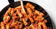 a skillet filled with pasta and meat on top of a white countertop next to a wooden spoon