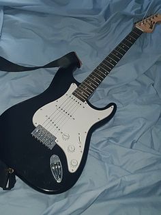 an electric guitar laying on top of a blue cloth covered bed sheet with a black strap