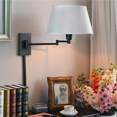 a lamp on a wall next to a table with books and a vase filled with flowers