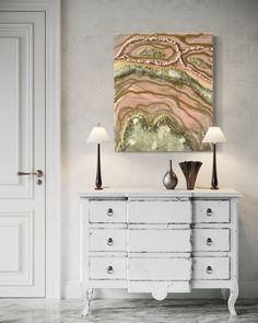 an abstract painting hangs on the wall above a white dresser with two lamps in front of it